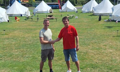 2 Männer schütteln sich die Hände. Im Hintergrund sind die Zelte des Sommercamps zu sehen.